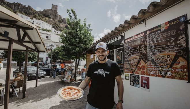 Enrico Granieri muestra una de sus pizzas artesanas, con su quiosco al fondo y el castillo de Zahara de la Sierra, donde ha echado raíces junto a su familia, en lo más alto.