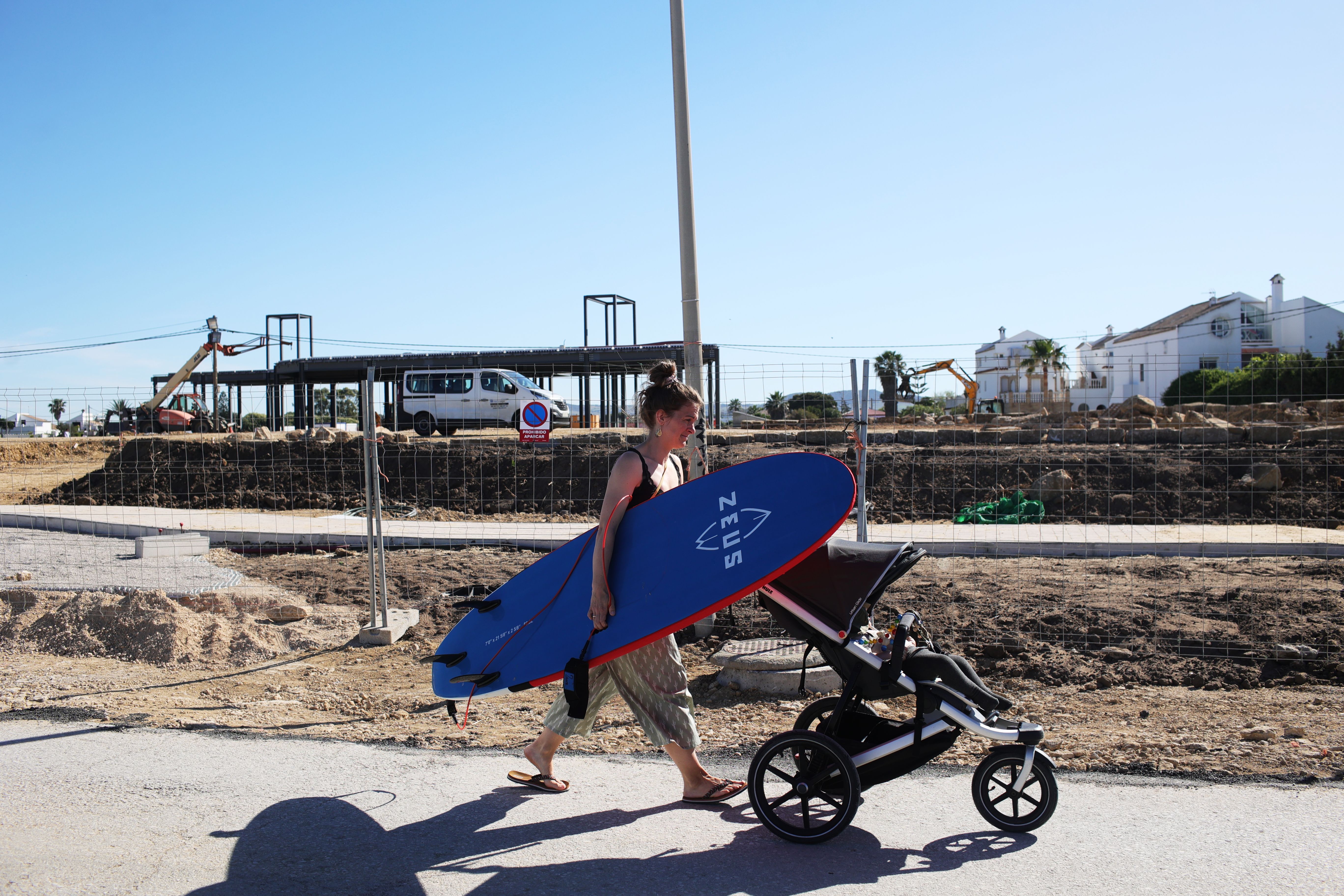 Una mujer pasa con una tabla de surf y un bebé por delante de la obra del 'beach club' Valhalla, en El Palmar. Una mujer pasa con una tabla de surf y un bebé por delante de la obra del 'beach club' Valhalla, en El Palmar.