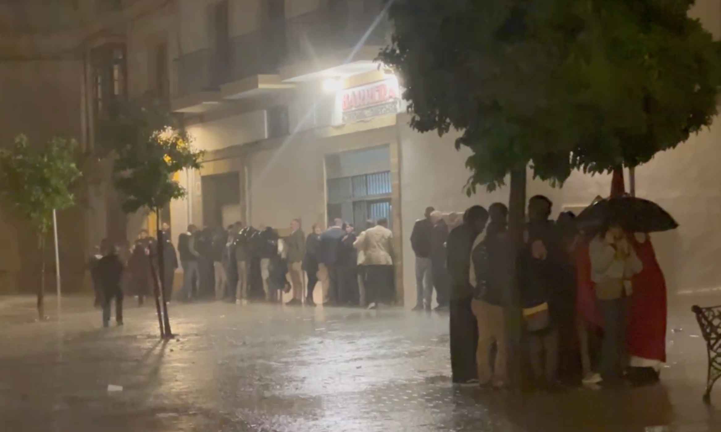 Un buen número de personas trata de protegerse de la lluvia, con nazarenos incluidos bajo un árbol.