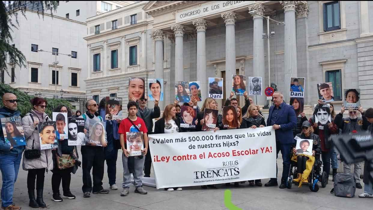 Integrantes de Trencats y familiares de víctimas posan frente al Congreso de los Diputados sosteniendo retratos de sus hijas e hijos.